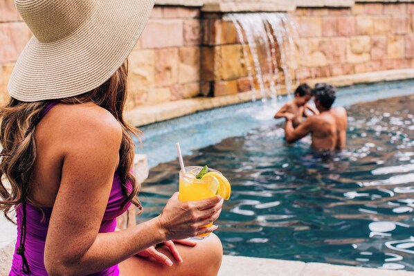 A woman in a purple swimsuit holding a drink by a pool with a waterfall in the background.
