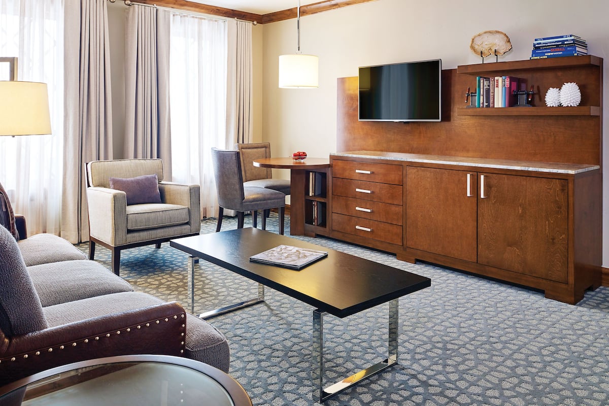 A hotel room with a sofa, armchair, dining table, TV, wooden cabinet, and bookshelves.