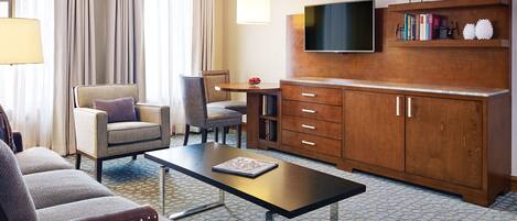 A hotel room with a sofa, armchair, dining table, TV, wooden cabinet, and bookshelves.