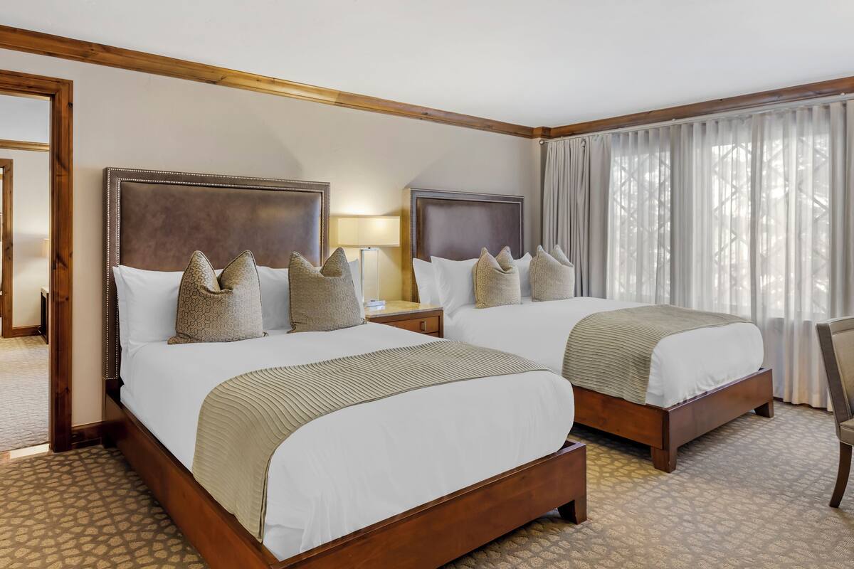 A hotel room with two beds, wooden headboards, patterned pillows, and a view of a building through sheer curtains.