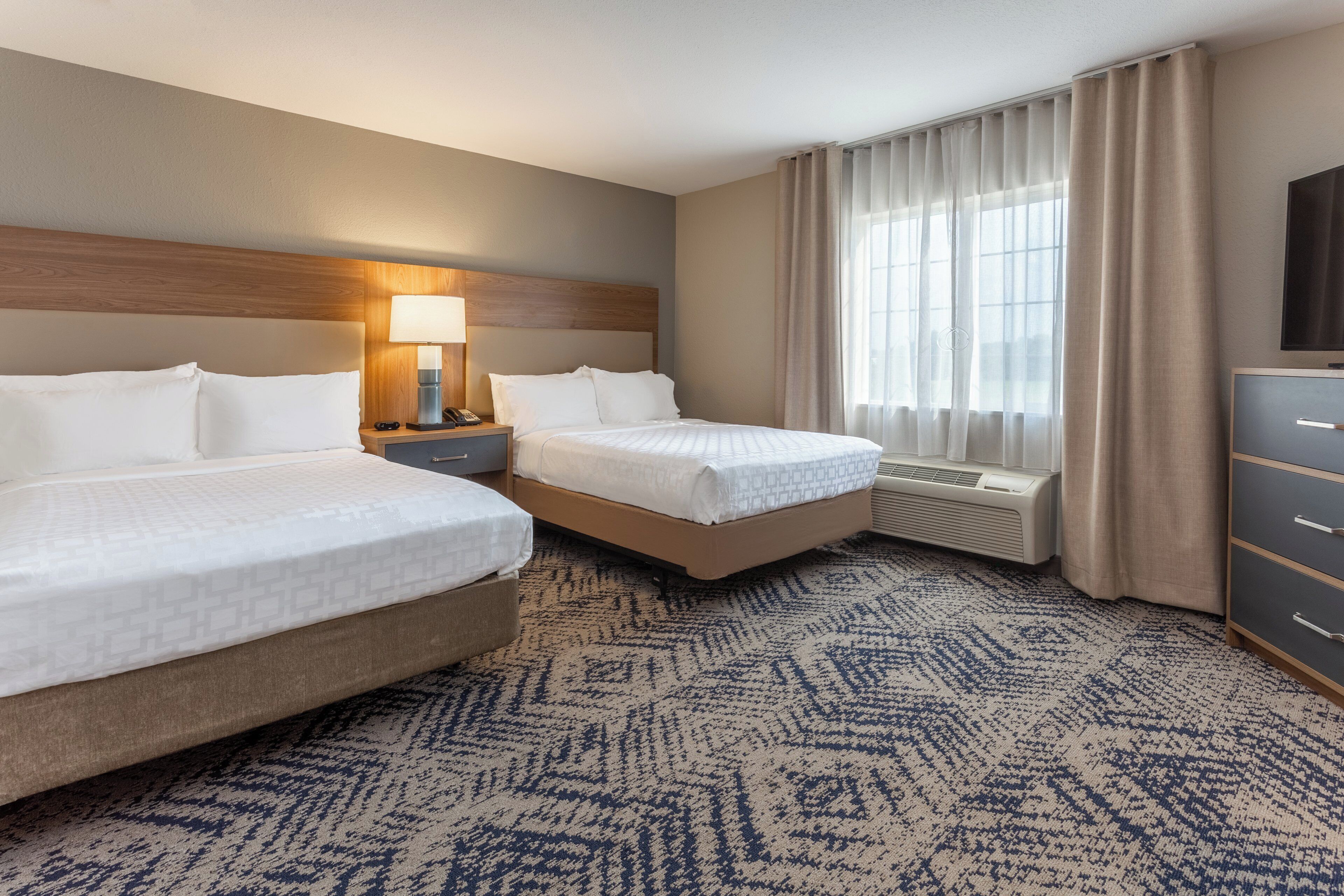 A hotel room with two beds, a desk with a lamp, a dresser, and a large window with curtains.