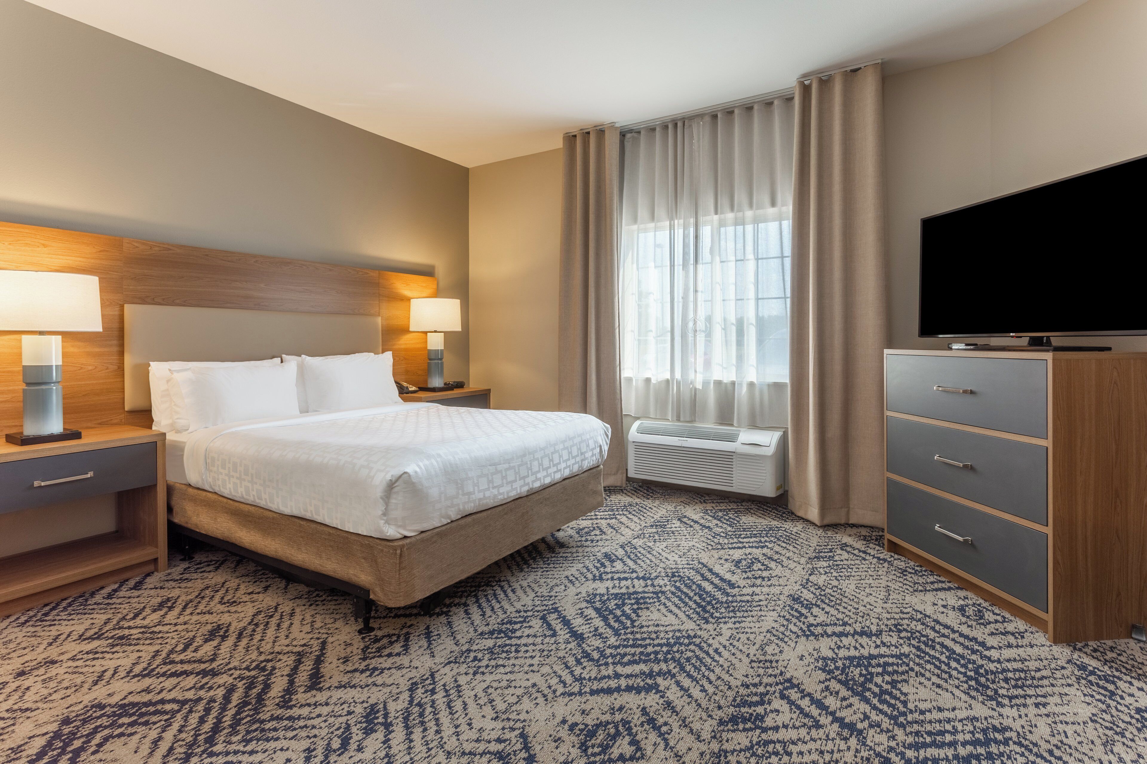 A hotel room with a bed, bedside lamps, a TV, a dresser, and a window with curtains.