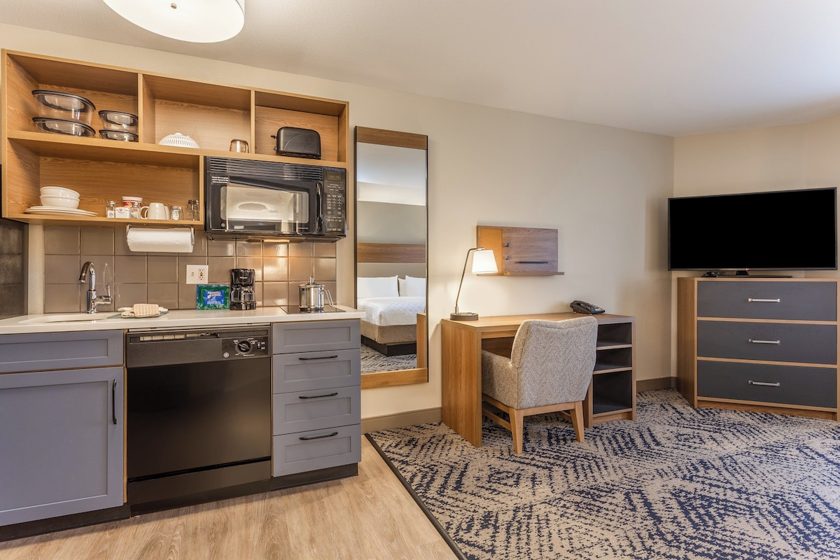 A compact hotel room with a kitchenette, a bed, a desk with a chair, a TV, and a wardrobe.