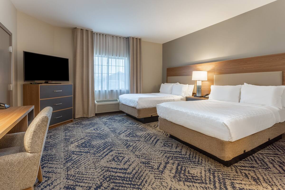 A hotel room with two beds, a desk, a television, and a large window with curtains.