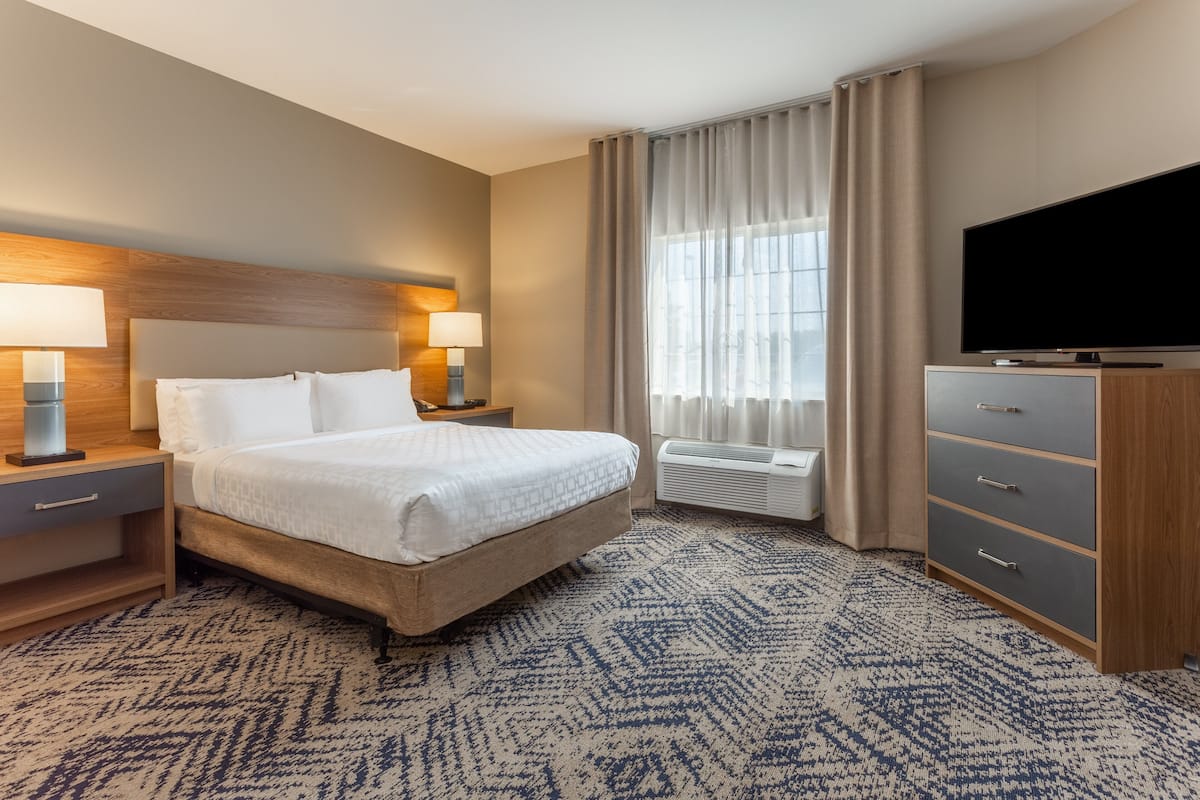 A hotel room with a large bed, two bedside tables with lamps, a flat-screen TV, a dresser, and a large window with curtains.