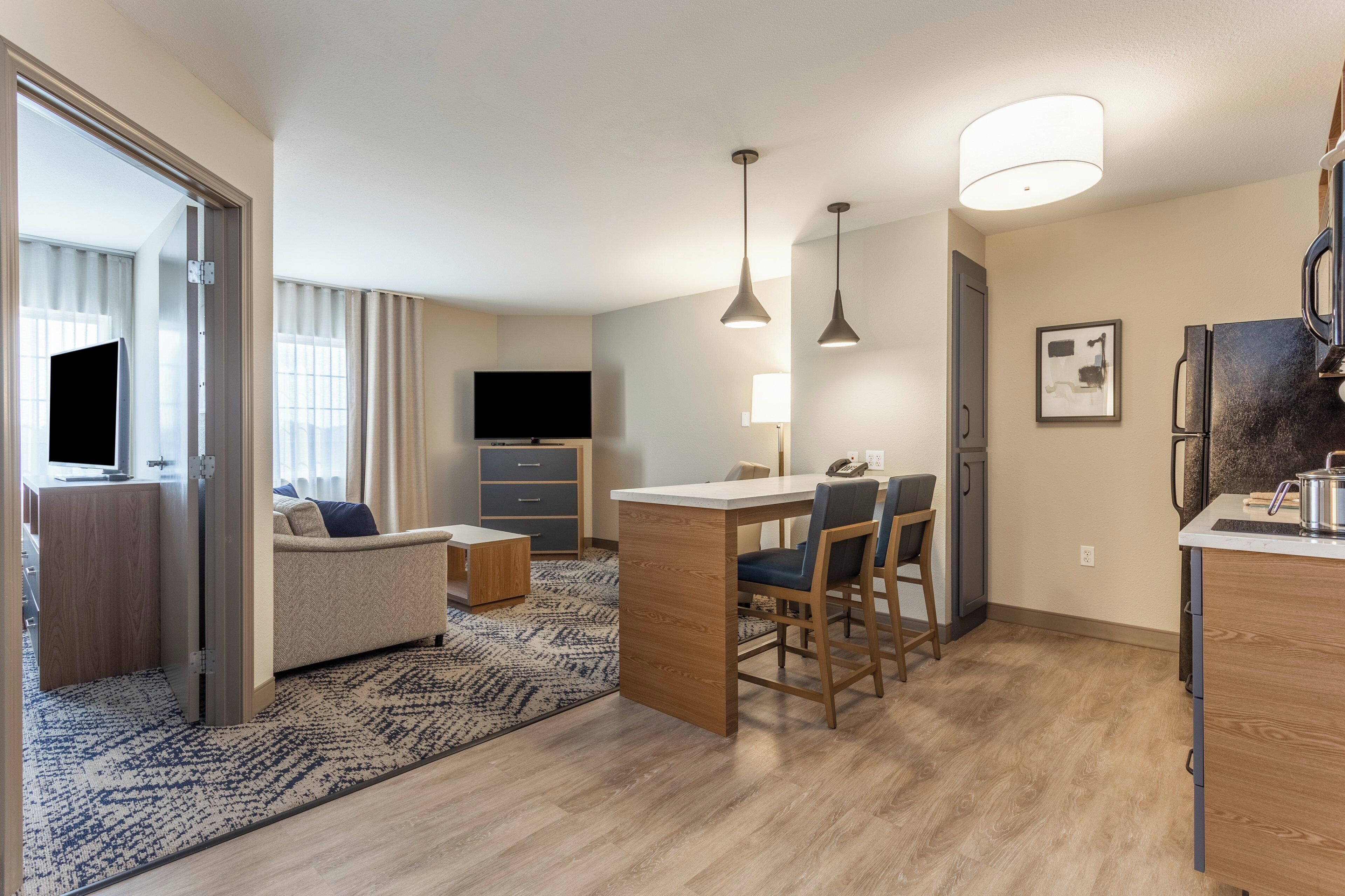 A modern hotel room with a flat-screen TV, a sofa, a dining area with chairs, a kitchen counter, and a large window with curtains.