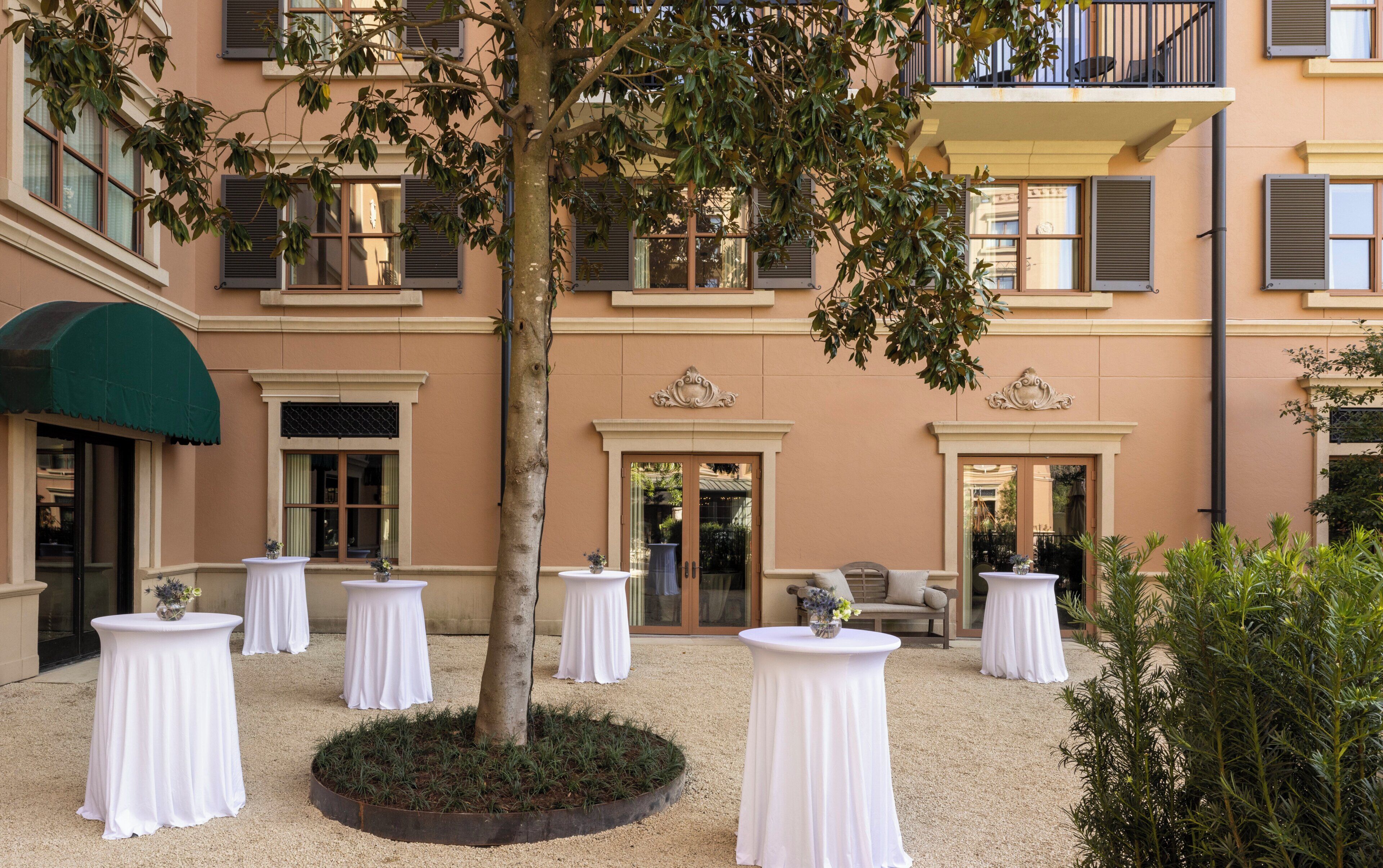 Une cour avec des tables et des chaises blanches, un arbre et la façade d’un immeuble agrémentée de balcons et de fenêtres.