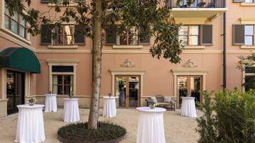 Une cour avec des tables et des chaises blanches, un arbre et la façade d’un immeuble agrémentée de balcons et de fenêtres.