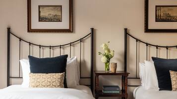 Une chambre d’hôtel avec deux lits, chacun agrémenté de draps blancs et d’oreillers foncés, une table de chevet avec un livre et un vase de fleurs, et deux tableaux encadrés accrochés au mur.