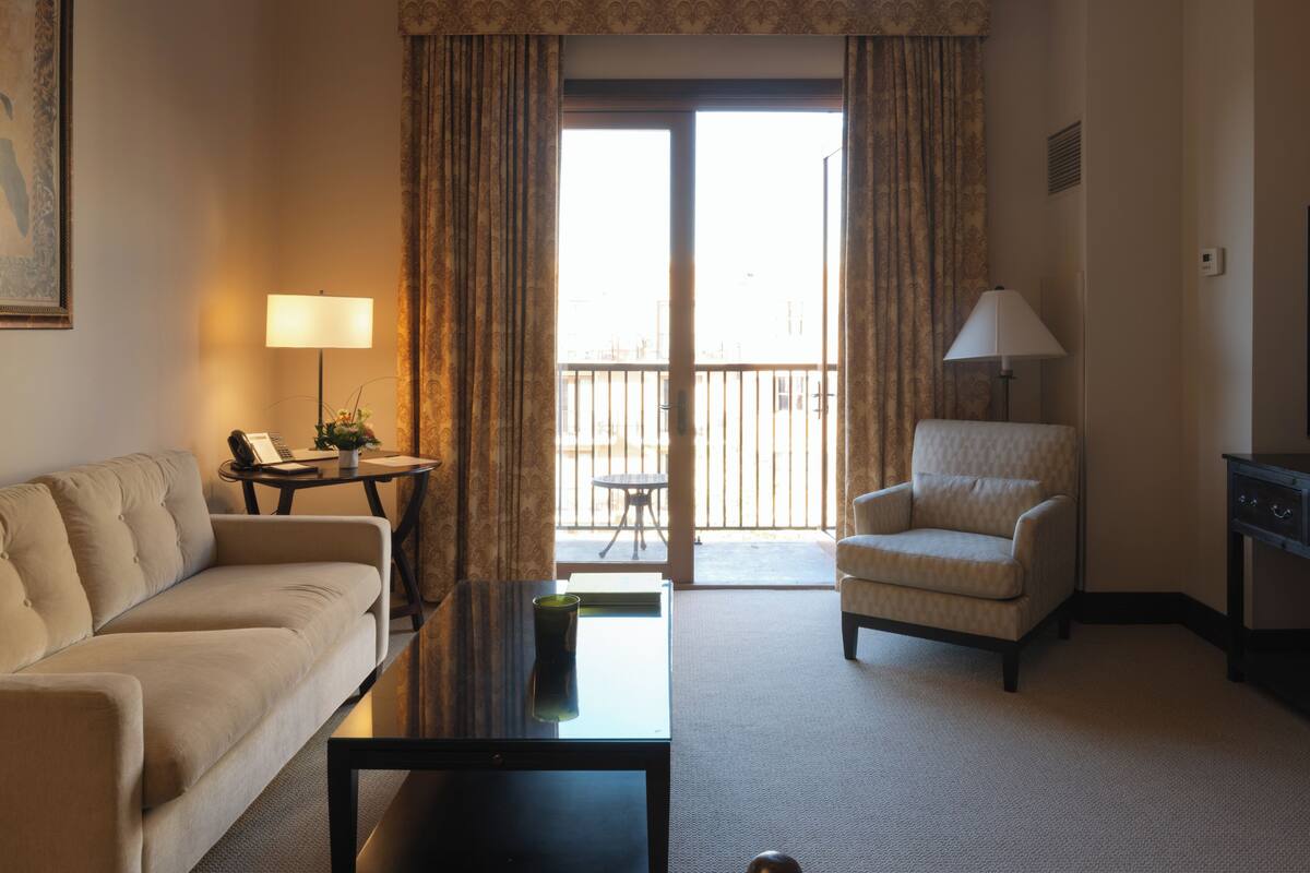 A hotel room with a sofa, armchair, coffee table, lamp, and a view through a large window.
