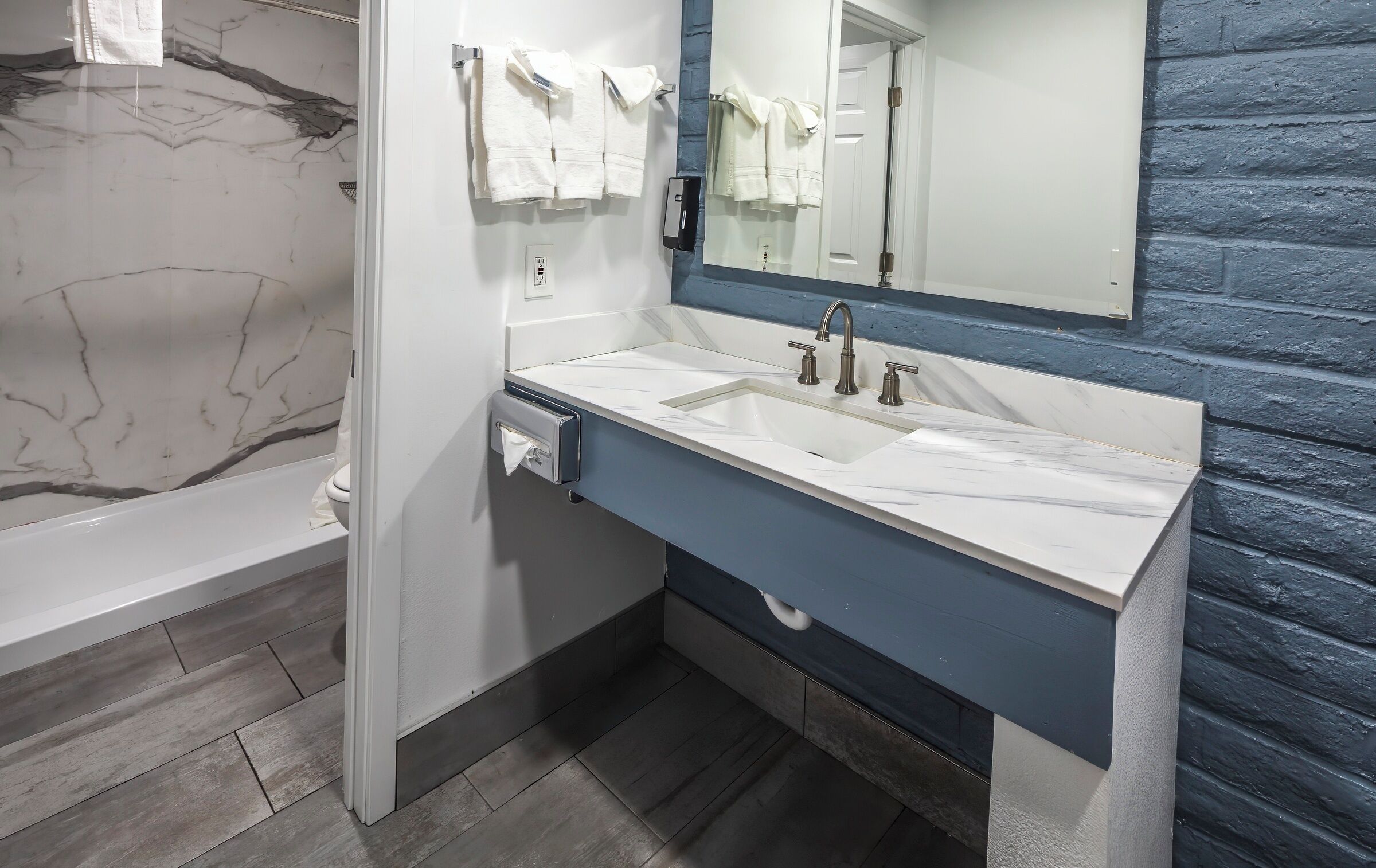A bathroom with a marble-tiled wall, a white bathtub, a sink with a blue countertop, and a mirror above the sink.