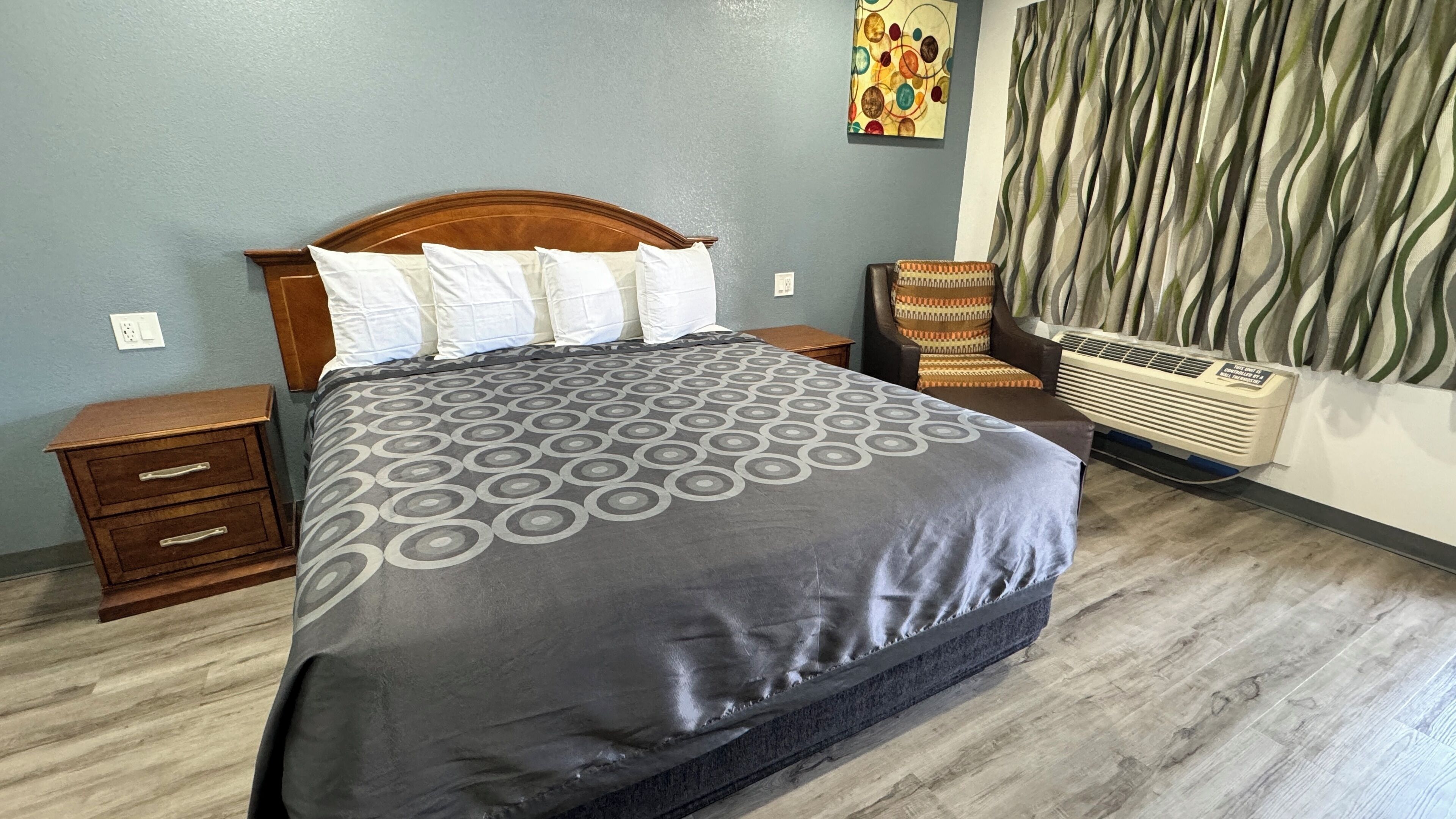 A neatly made bed with a patterned bedspread, a wooden nightstand, a chair with a patterned cushion, and a wall-mounted air conditioning unit.