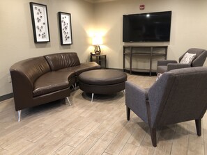 A living room with a brown leather sofa, a grey armchair, a round ottoman, a flat-screen TV, and two framed artworks on the wall. - Candlewood Suites Fort Wayne by IHG (Fort Wayne)