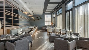 A modern restaurant with long tables, grey upholstered chairs, and large windows allowing natural light.