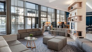 A modern hotel lobby with large windows, a seating area with sofas and ottomans, a small coffee table, and a bookshelf with various items.