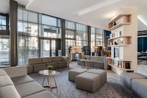 Un moderno hall de hotel con ventanas amplias, un área de descanso con sofás y otomanas, una mesita de centro y una estantería con diversos objetos.