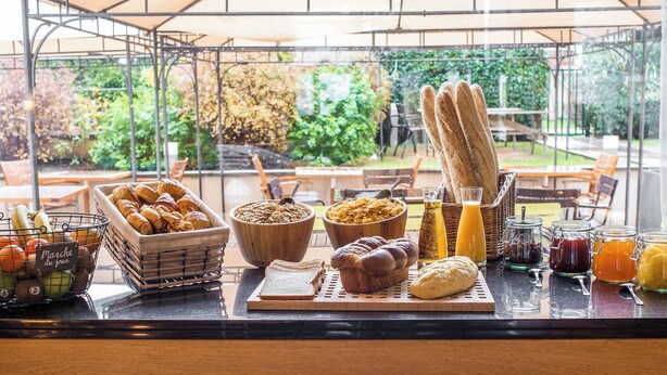 A breakfast buffet with bread, pastries, fruits, and beverages on a table under a canopy.