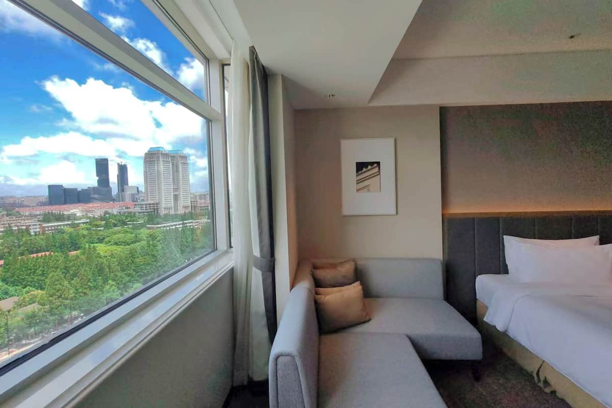Habitación de hotel con un ventanal amplio que ofrece vistas a la ciudad, un sofá gris, una cama y un cuadro en la pared.