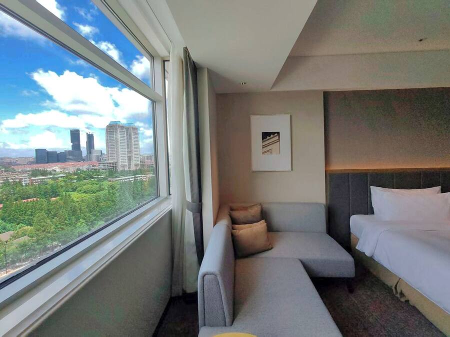Ein Hotelzimmer mit großem Fenster und Blick auf die Stadt, einem grauen Sofa, einem Bett und einem gerahmten Bild an der Wand.