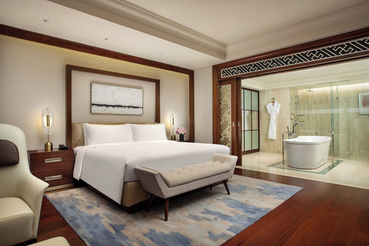 Une chambre d’hôtel avec un grand lit, des tables de chevet, un banc, un tapis et une salle de bain équipée d’une baignoire et d’une douche.