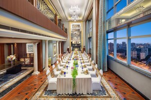 A banquet hall with a long table set for a formal event, large windows with a city view, and a chandelier.