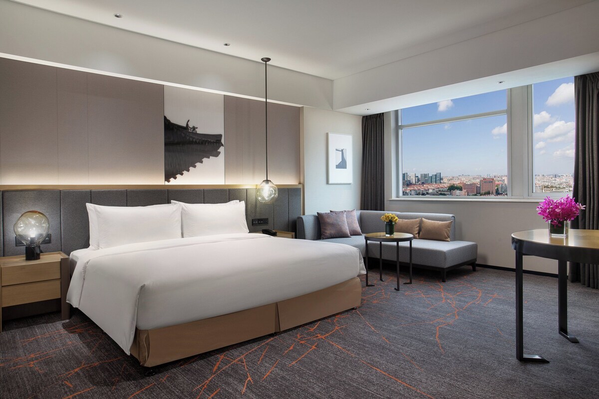 Une chambre d’hôtel moderne, dotée d’un grand lit, d’un canapé, d’une petite table et offrant une vue sur la ville par la fenêtre.
