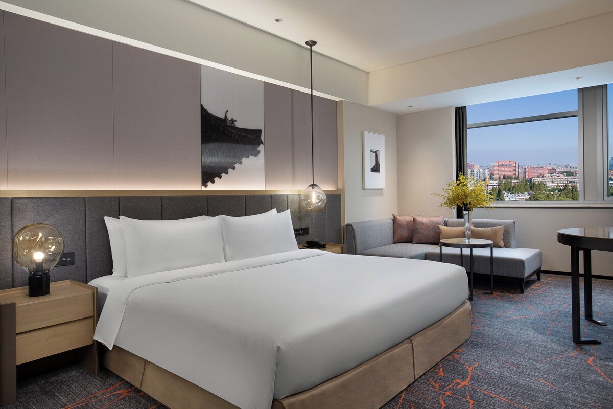 Une chambre d’hôtel moderne, dotée d’un grand lit, d’un bureau, d’une chaise et offrant une vue sur la ville par la fenêtre.