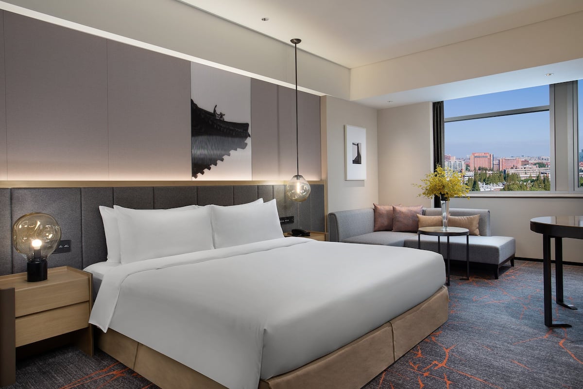 Una habitación de hotel moderna con una cama grande, un escritorio, una silla y vistas a la ciudad a través de un ventanal amplio.