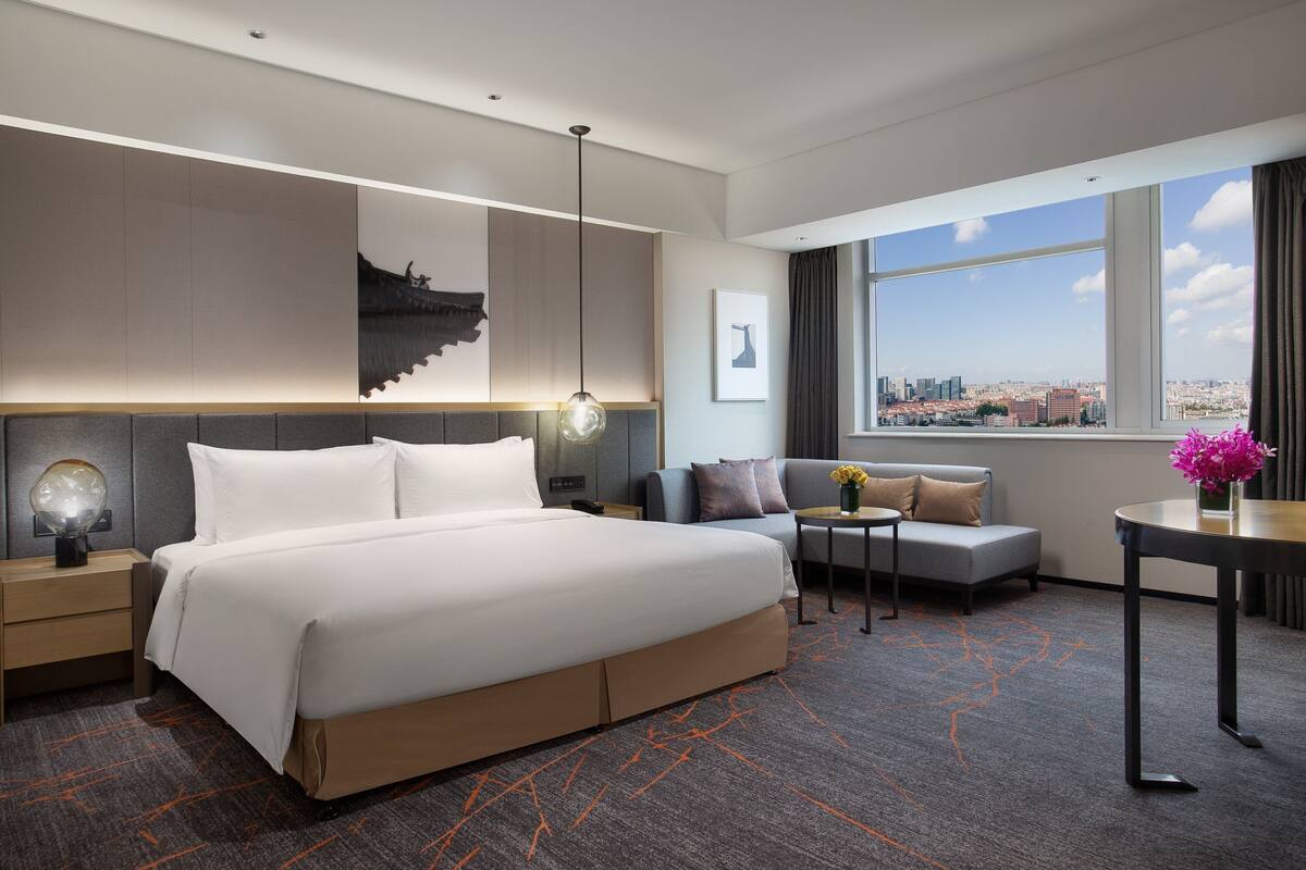 Una habitación de hotel moderna con una cama grande, un sofá, una mesita con flores y vistas a la ciudad a través de amplias ventanas.