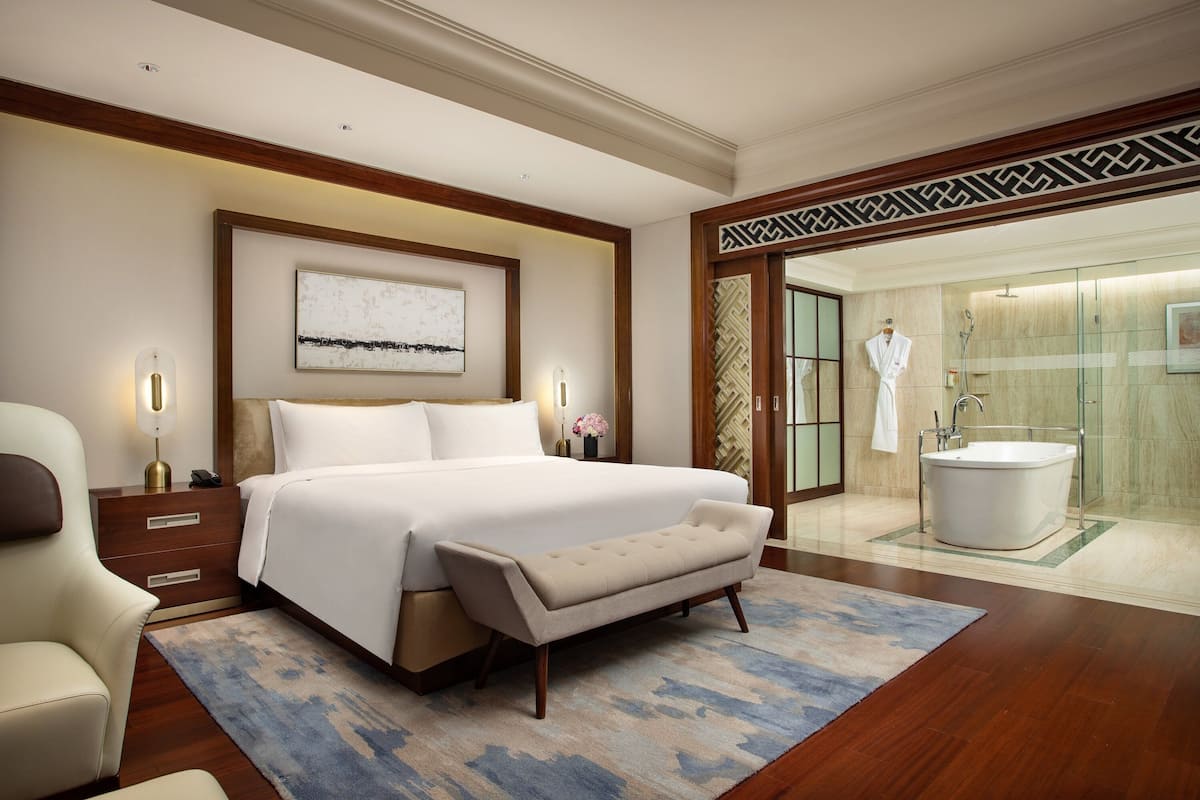 Una habitación de hotel con una cama grande, mesitas de noche, un banco, un baño con bañera y cabina de ducha de vidrio.