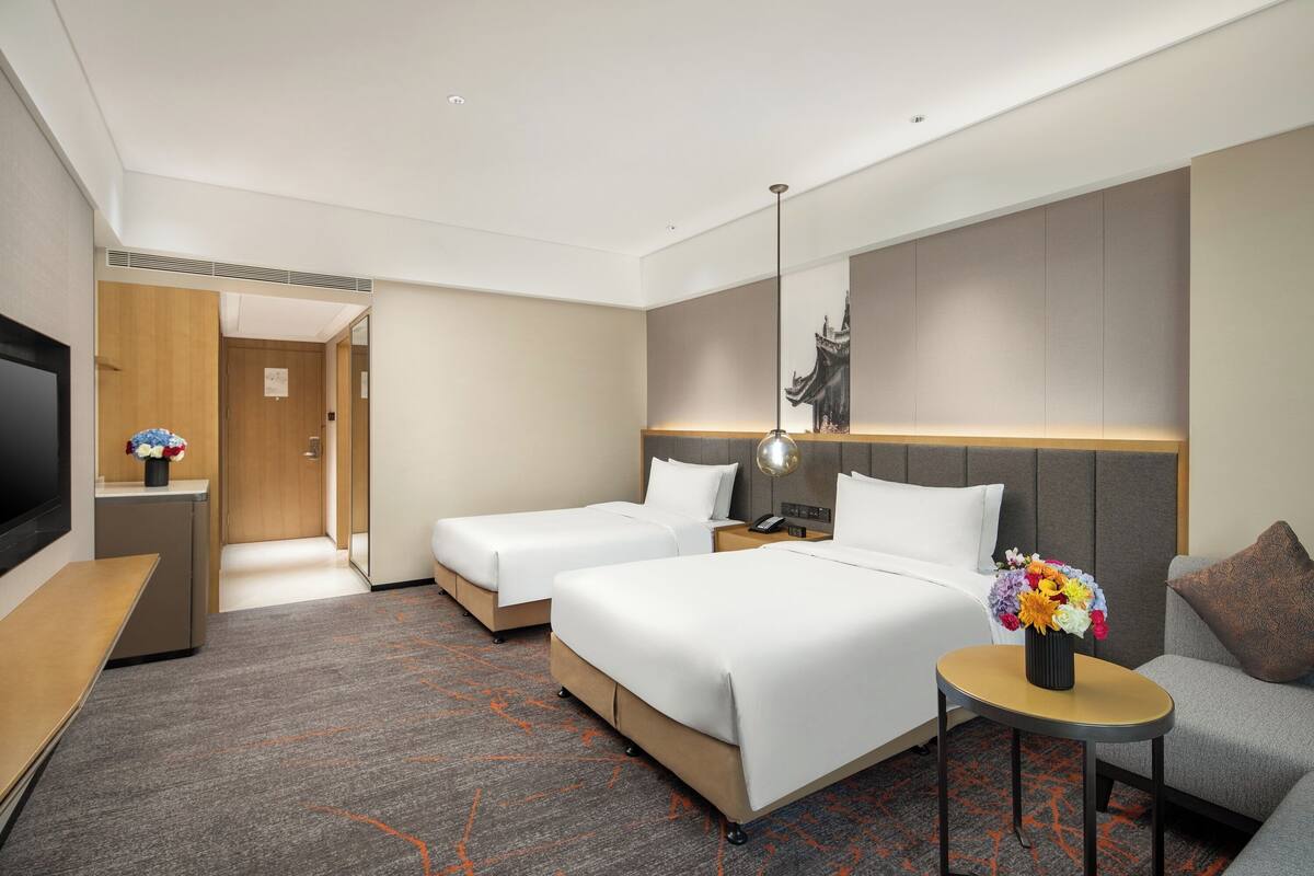 Habitación de hotel con dos camas, un escritorio, un televisor, una mesita con un florero y un sofá.