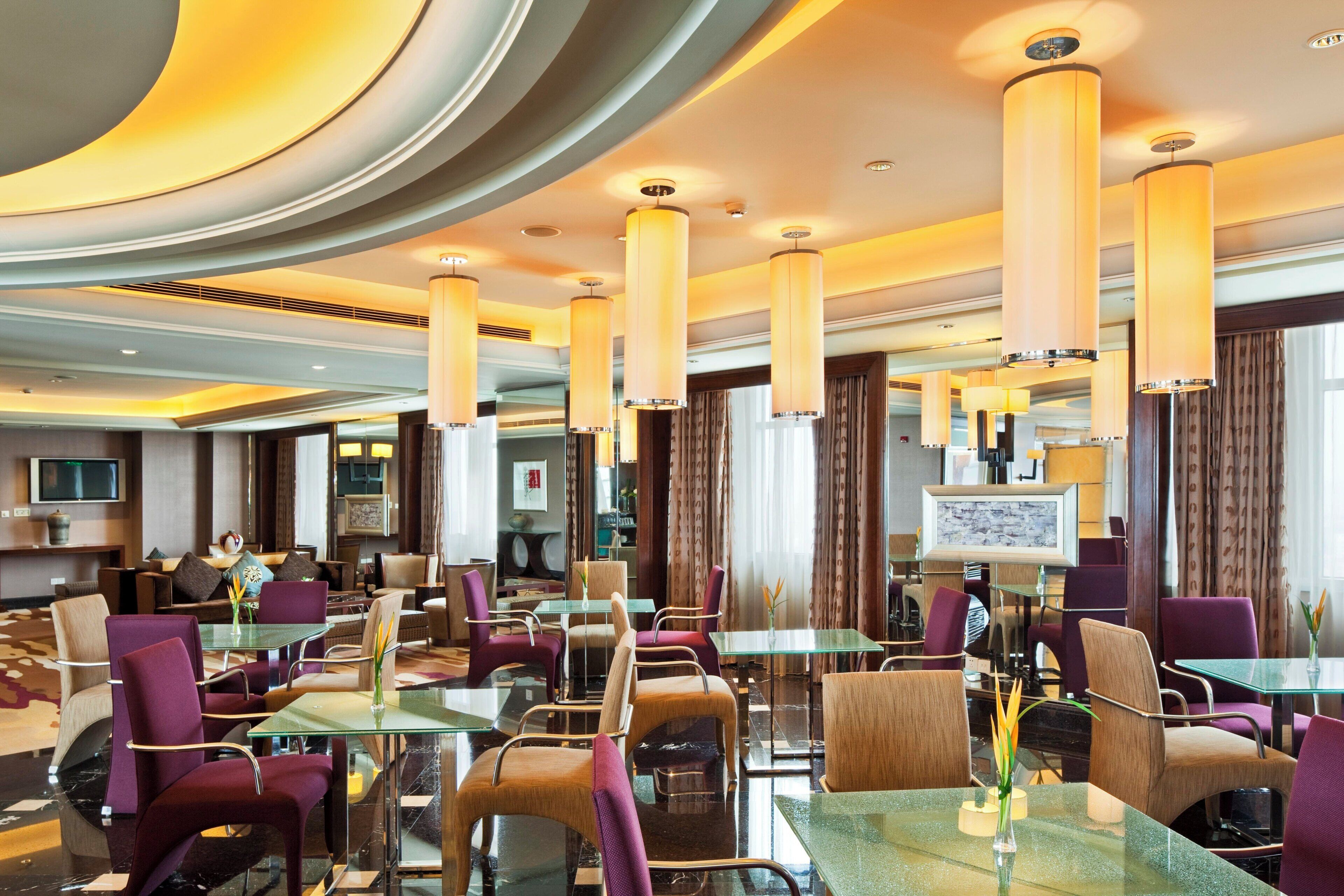 Un hall d’hôtel doté d’un mobilier moderne, comprenant des tables à plateaux en verre et des fauteuils violets, ainsi qu’un grand écran de télévision.