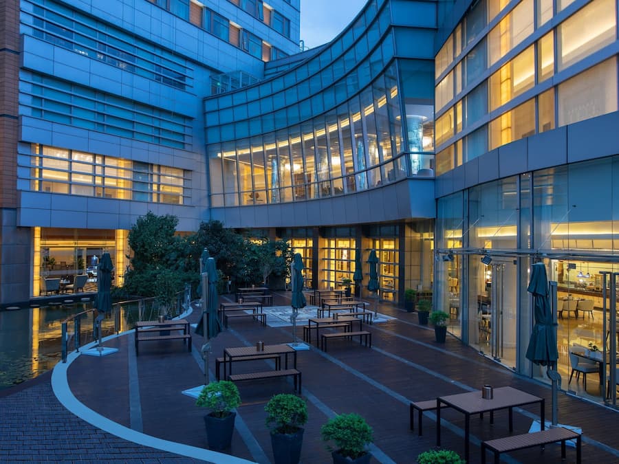 A modern building with a curved glass facade, outdoor seating area with tables and umbrellas, and a pool in the foreground.