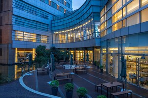 A modern building with a curved glass facade, outdoor seating area with tables and umbrellas, and a pool in the foreground.