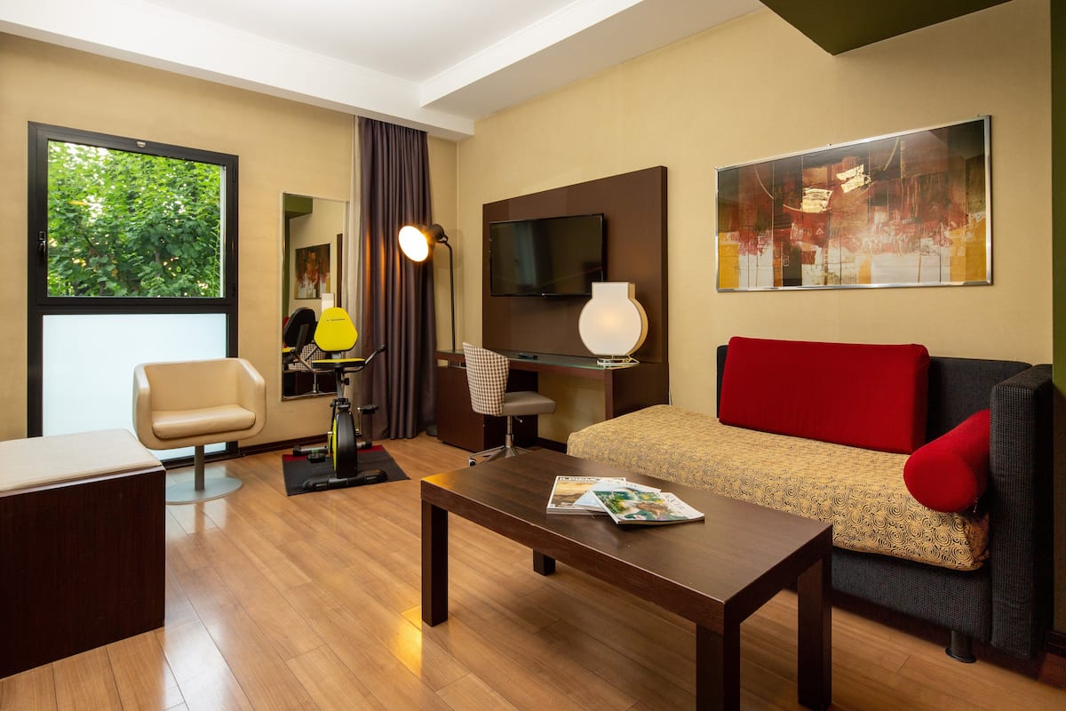 A modern hotel room with a wooden floor, a sofa with a red cushion, a desk with a lamp, a TV, and a large window with a view of trees.