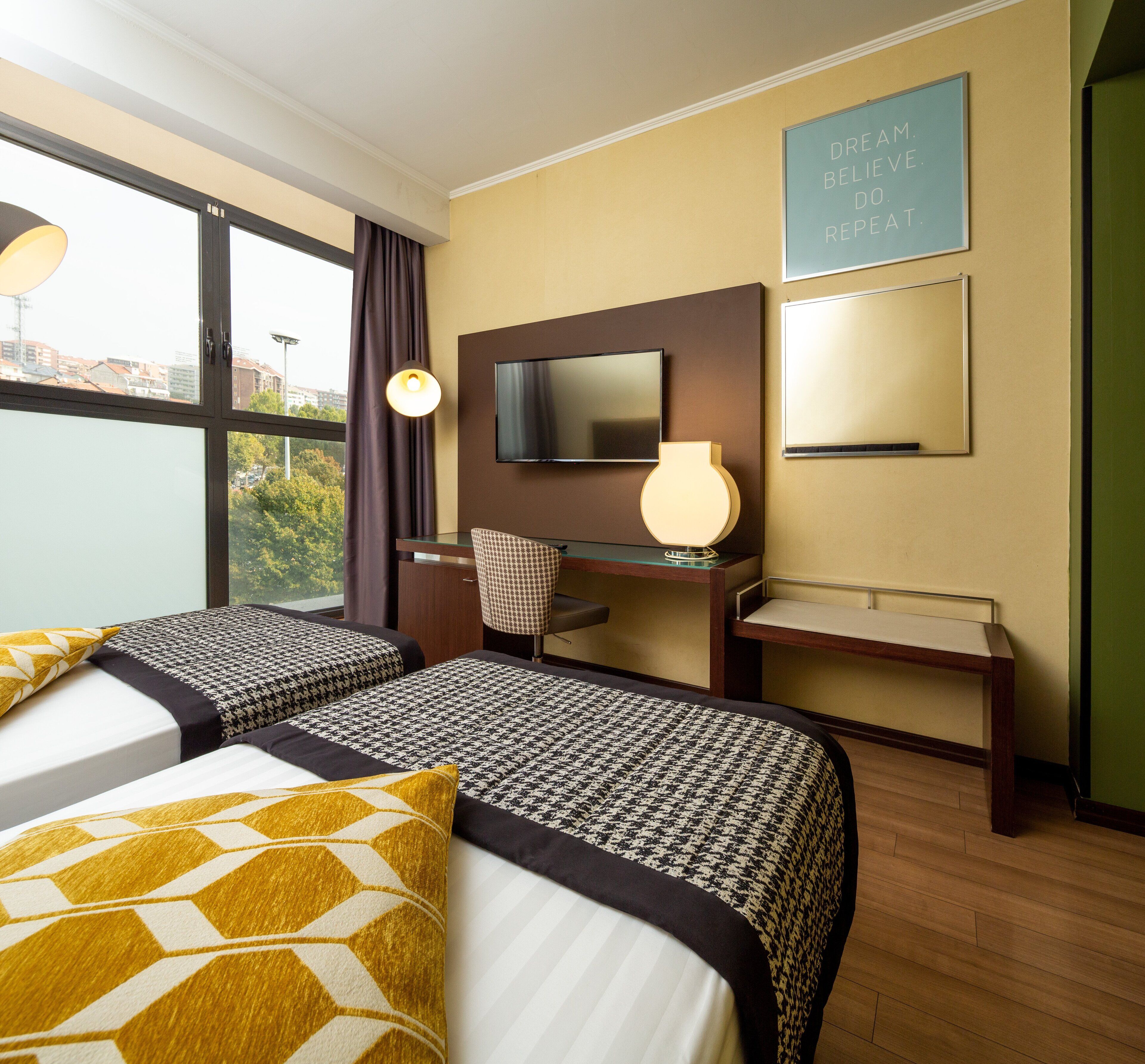 A hotel room with two beds, a desk with a chair, a television, and a motivational quote on the wall.