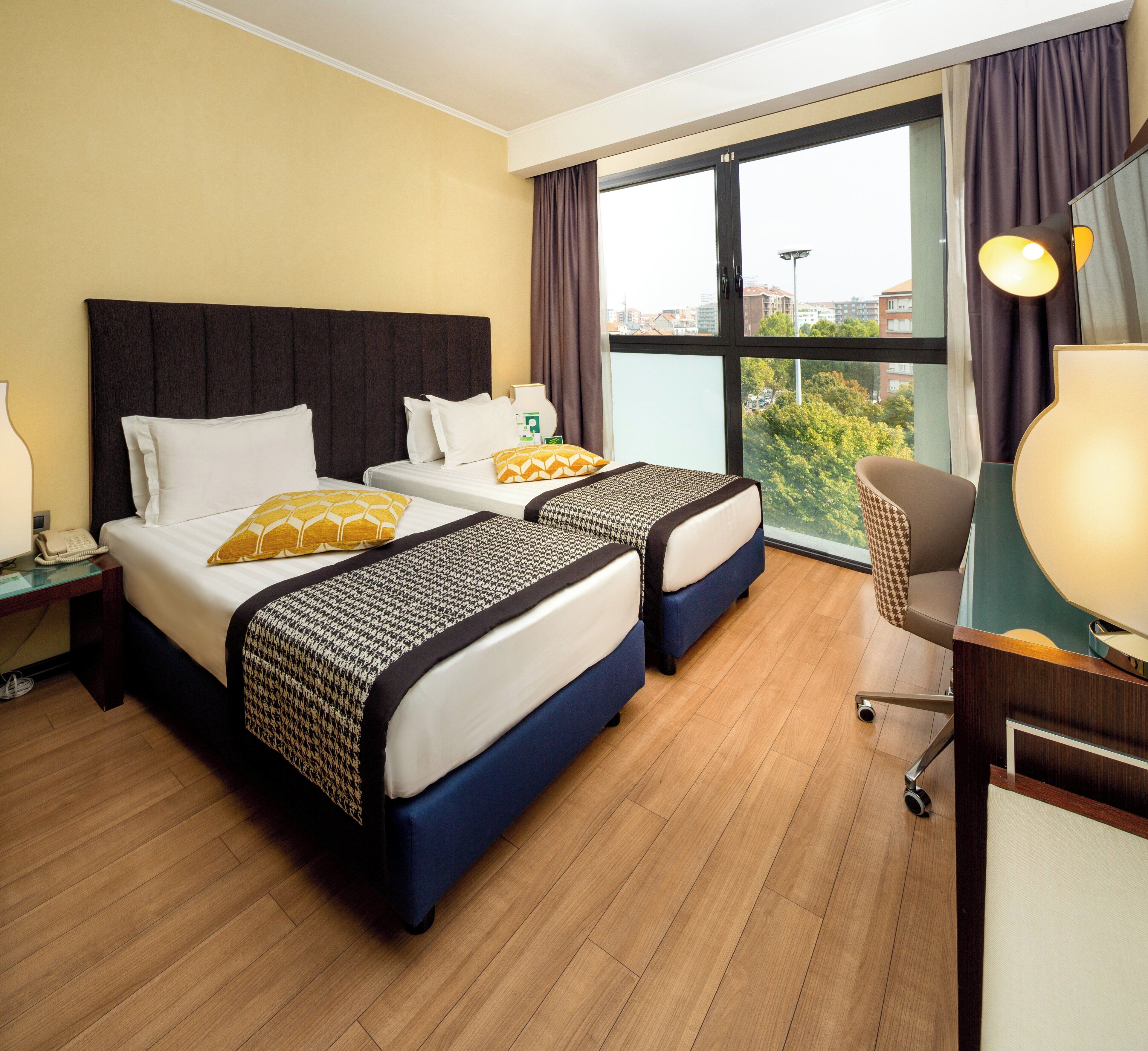 A hotel room with two beds, a desk, and a view of the city.