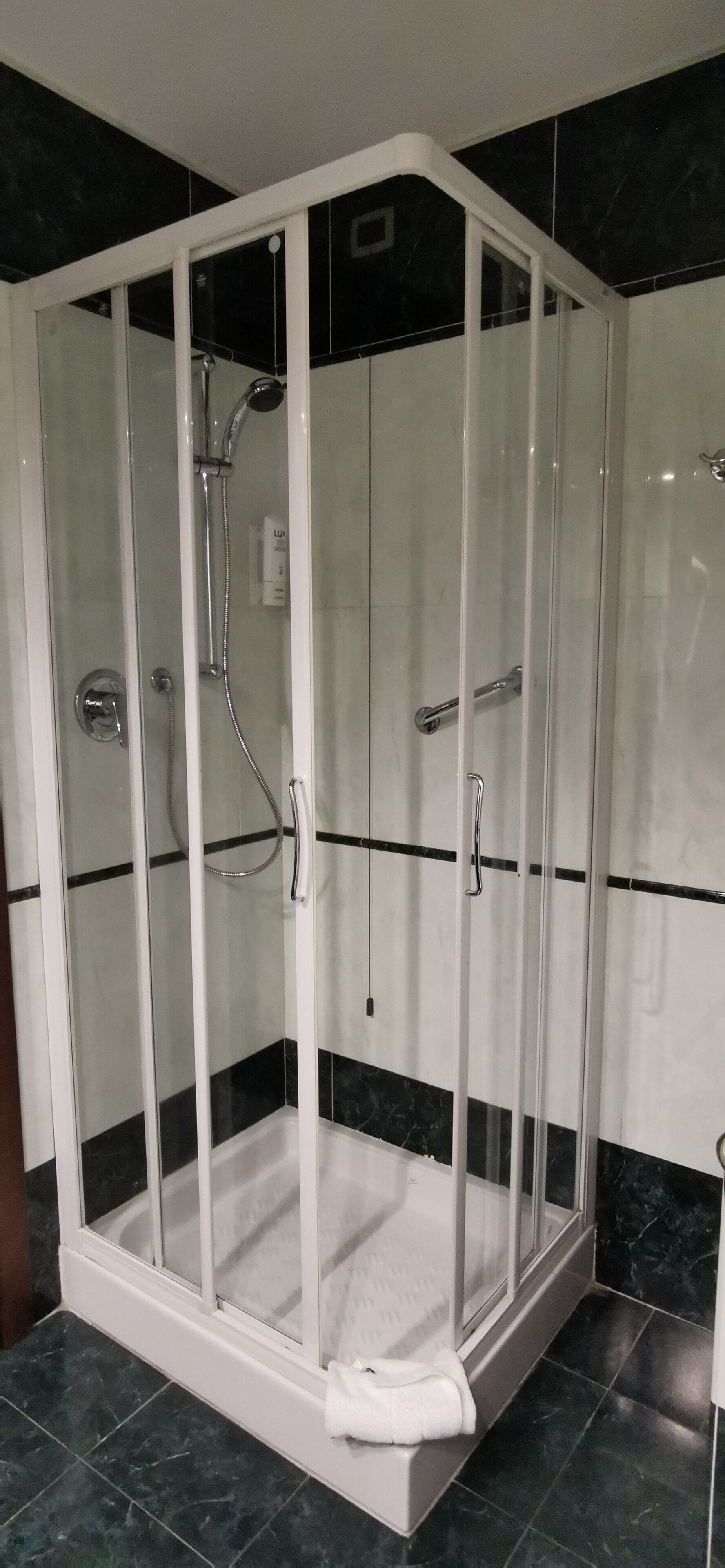 A glass-enclosed shower area with a white base, black and white tiled walls, and a white towel on the floor.