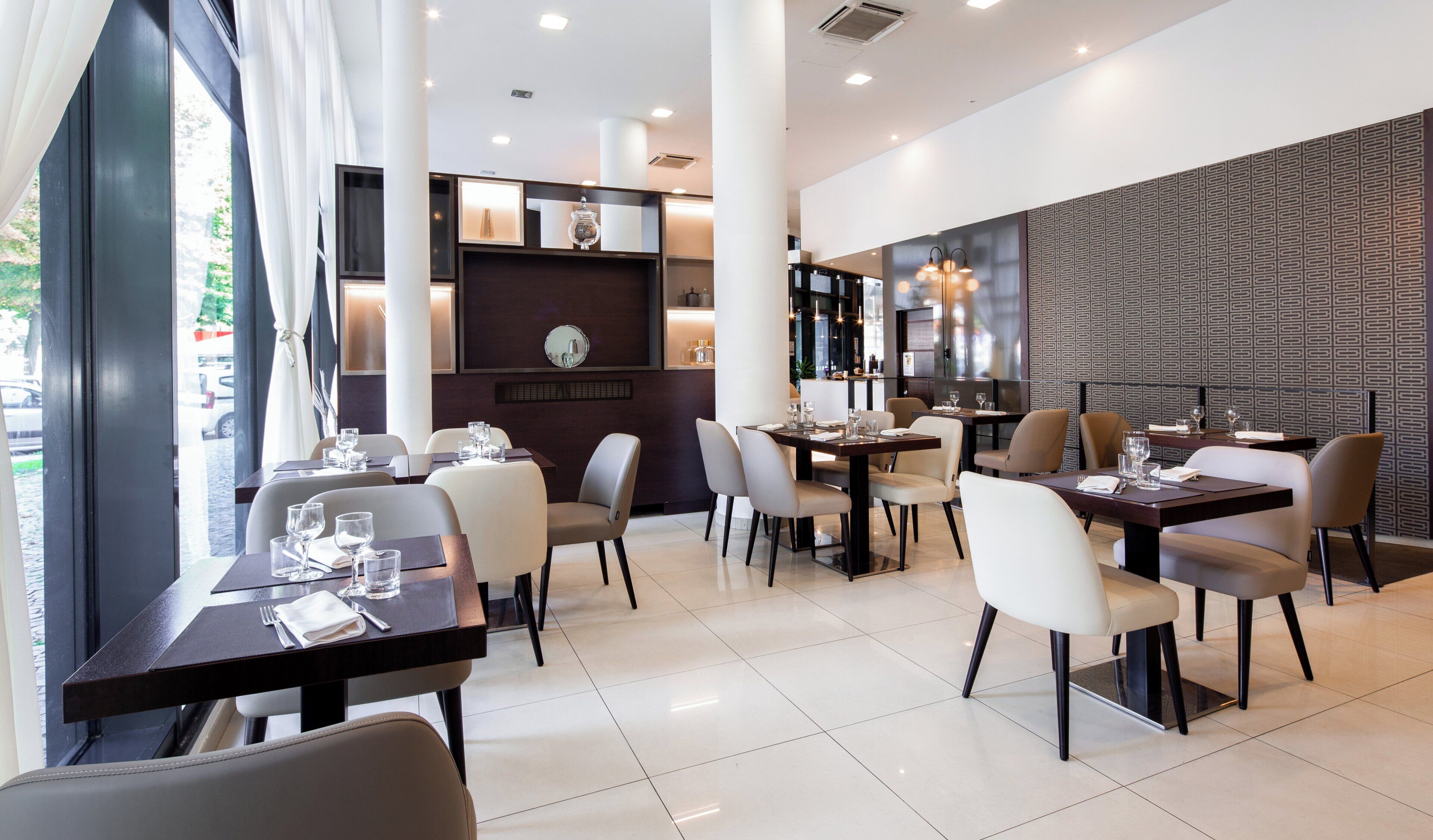 A modern restaurant interior with multiple dining tables and chairs, a patterned wall, and a view of the street outside.