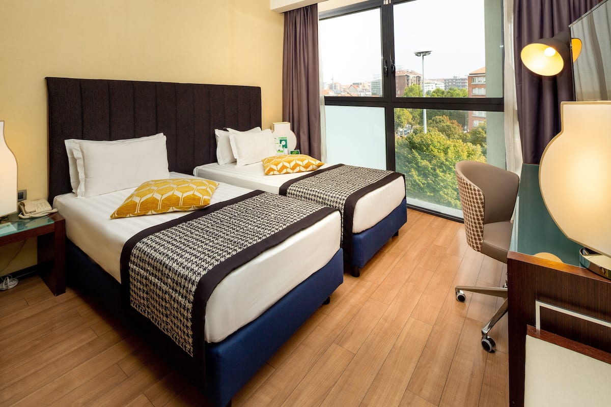 A hotel room with two beds, a desk, and a large window with a city view.