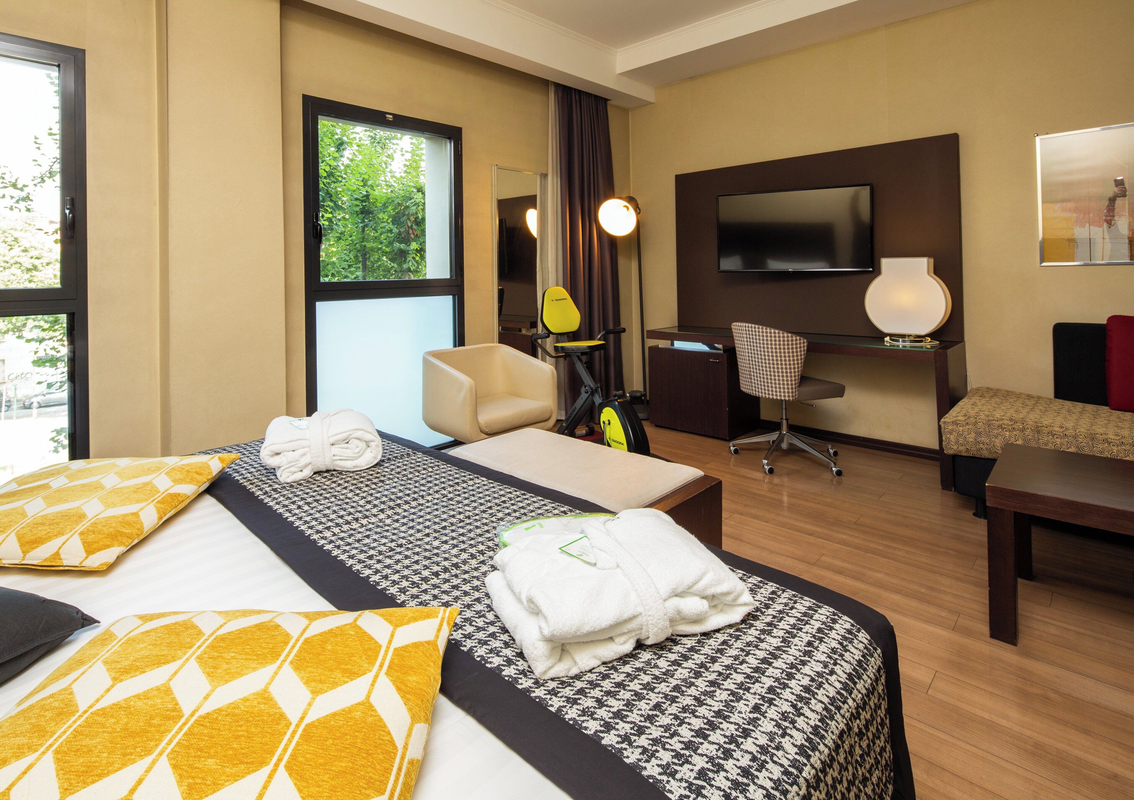 A hotel room with two beds, a desk, a chair, a TV, and a window with a view of greenery.