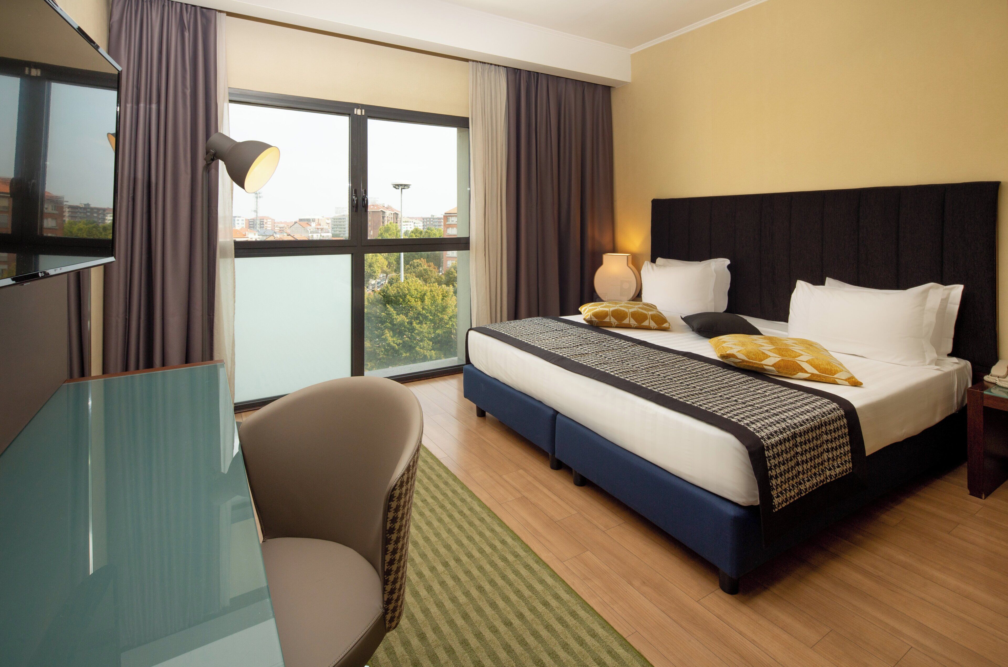 A hotel room with a bed, a desk, a chair, and a view of the city.