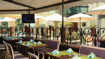 An outdoor cafe with tables and chairs, umbrellas, and a television screen.