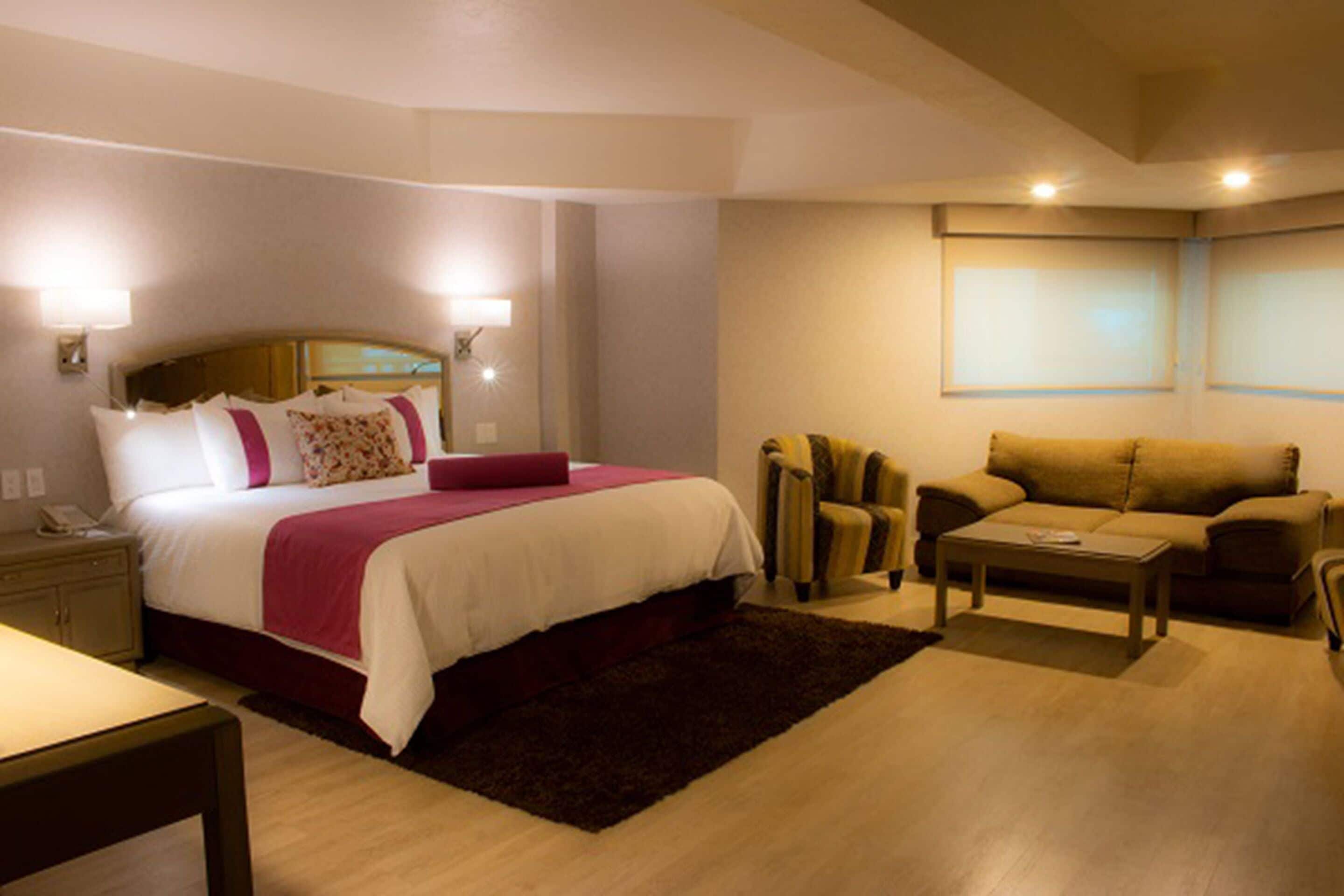 A hotel room with a bed, bedside table, lamp, sofa, and armchair.