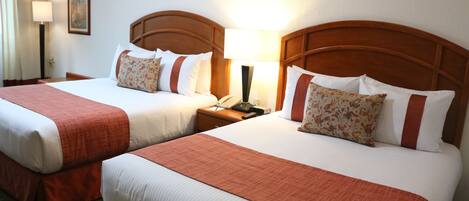 A hotel room with two beds, each with a wooden headboard, white bedding, and a red accent. There is a nightstand with a lamp and a telephone between the beds.