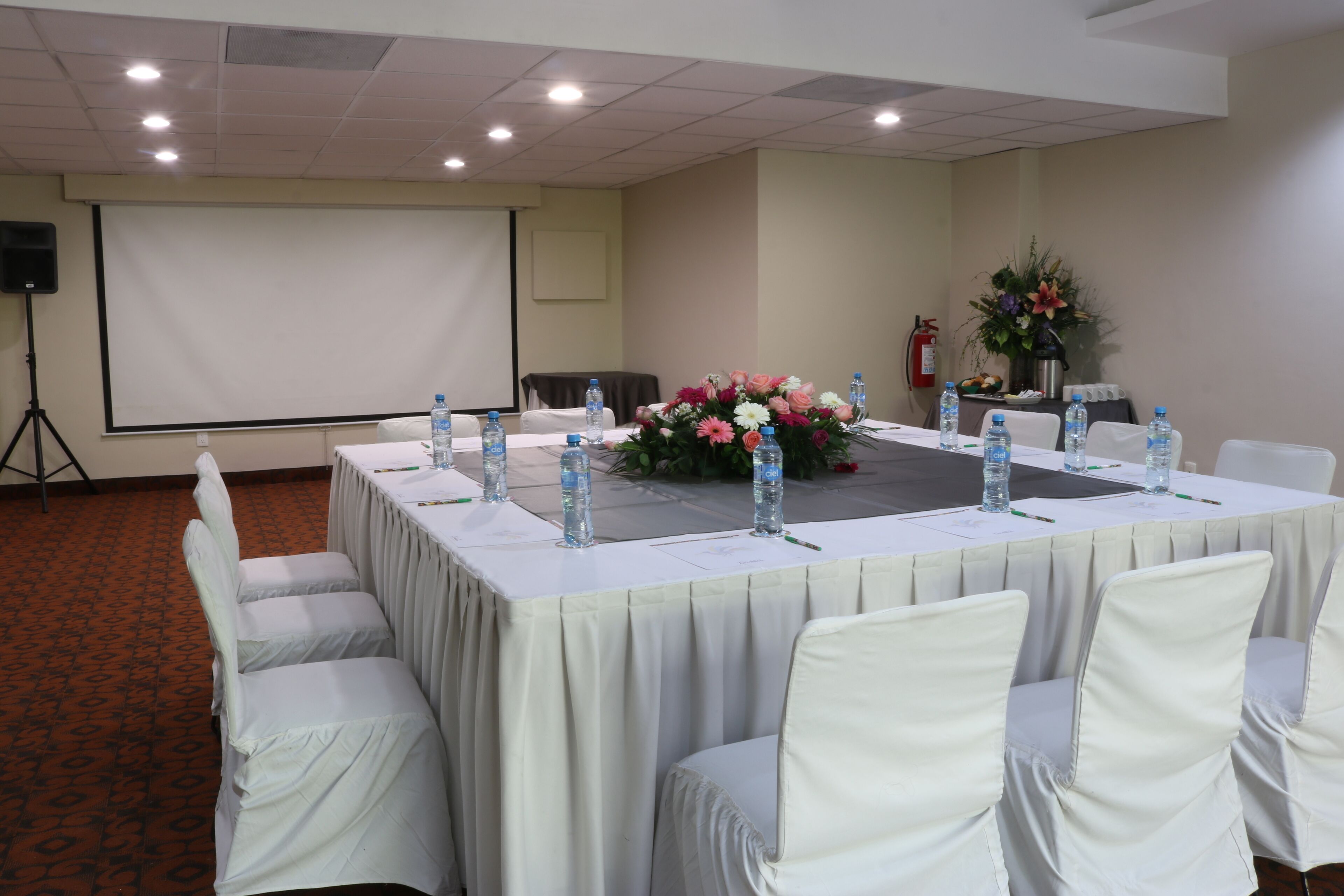 A conference room with a long table, white chairs, a projector screen, and a floral arrangement.