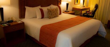 A hotel room with a large bed, a desk, a chair, a nightstand with a phone, and a lamp.