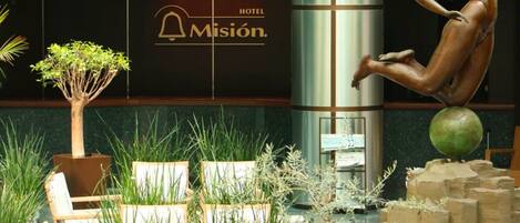 A hotel lobby with a bronze statue, a sign reading "HOTEL Misión", and indoor plants.