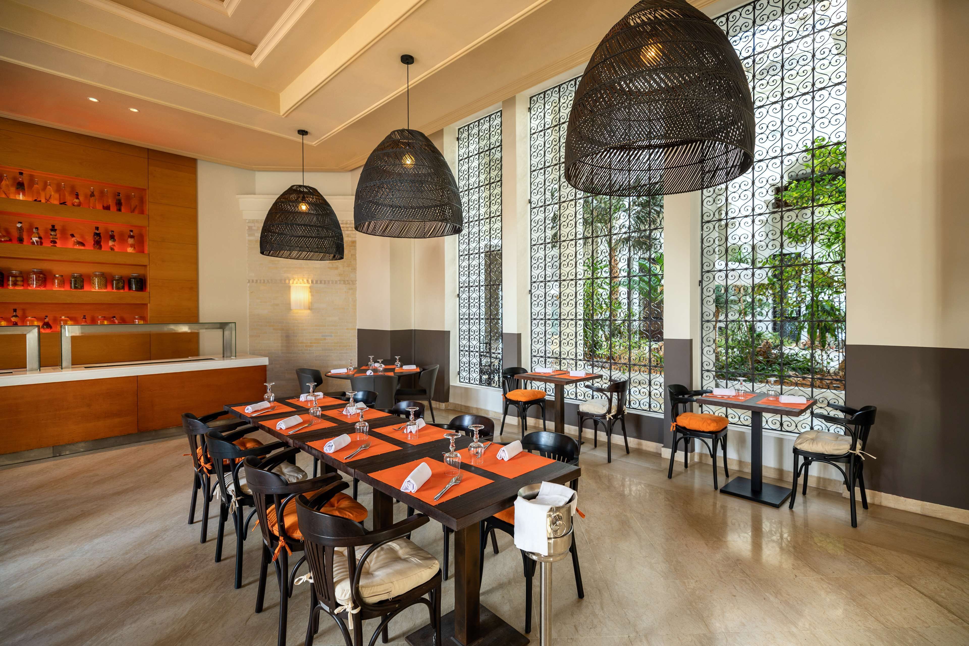 A restaurant with multiple tables and chairs, large windows with decorative iron grilles, and hanging black lamps.