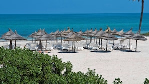 A beach with rows of white lounge chairs and thatched umbrellas, palm trees, and a clear blue sea. - Radisson Blu Athénée Palace Resort & Thalasso, Djerba (Mezraia)